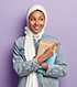 Isolated shot of satisfied Muslim student shows awesome advert, points at upper right corner, wears white veil and jean jacket, carries notepads , prepares for classes, isolated over purple wall